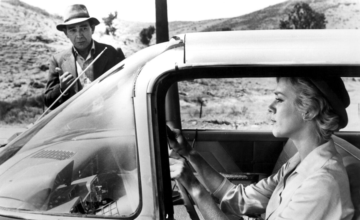black and white image from "The Hitch-Hiker" episode of "The Twilight Zone." In the foreground of the photo is a woman played by Inger Stevens seen through the window of the car she is driving. Standing in the background of the photo, near the passenger side of her car, is the mysterious hitchhiker, played by Leonard Strong, who has constantly been turning up along her route.