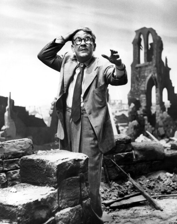 black and white photo from the "Time Enough at Last" episode of "The Twilight Zone." It depicts a character played by Burgess Meredith looking frightened as he stands in a post-apocalyptic world. He is standing amid rubble and ruined buildings, wearing a rumpled suit and eyeglasses.