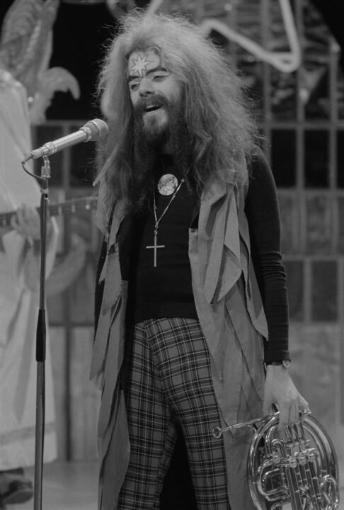 black-and-white image of Roy Wood, frontman for glam rock group Wizzard, performing on the Christmas Day 1973 broadcast of the BBC's "Top of the Pops." He has very long and shaggy hair and beard/mustaches, and is holding a French horn by his side in his left hand as he sings.