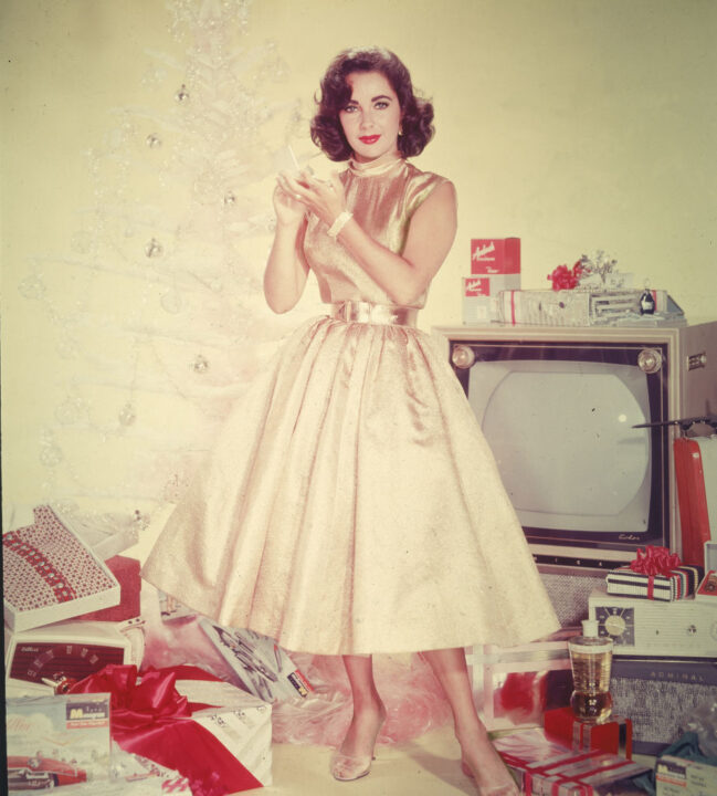 Publicity portrait of British-born actor Elizabeth Taylor in a yellow dress as she stands in front of an artificial Christmas tree amidst a pile of opened presents, including a television set, clothing, and a radio, circa 1950s 