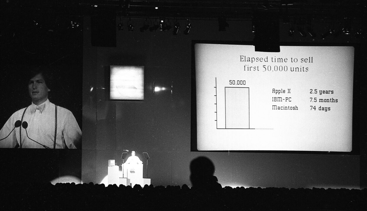 black and white image from April 23, 1984. Apple Computers chairman Steve Jobs is on a stage explaining the success of the company's new Macintosh computer, which had launched in late January. Jobs is very small on the stage, and to his left (audience's right) is a very large screen on which are some stats and charts comparing how long it took various computers to sell 50,000 units, with stacked text reading: "Apple II: 2.5 years" "IBM - PC: 7.5 months" "Macintosh: 74 days"