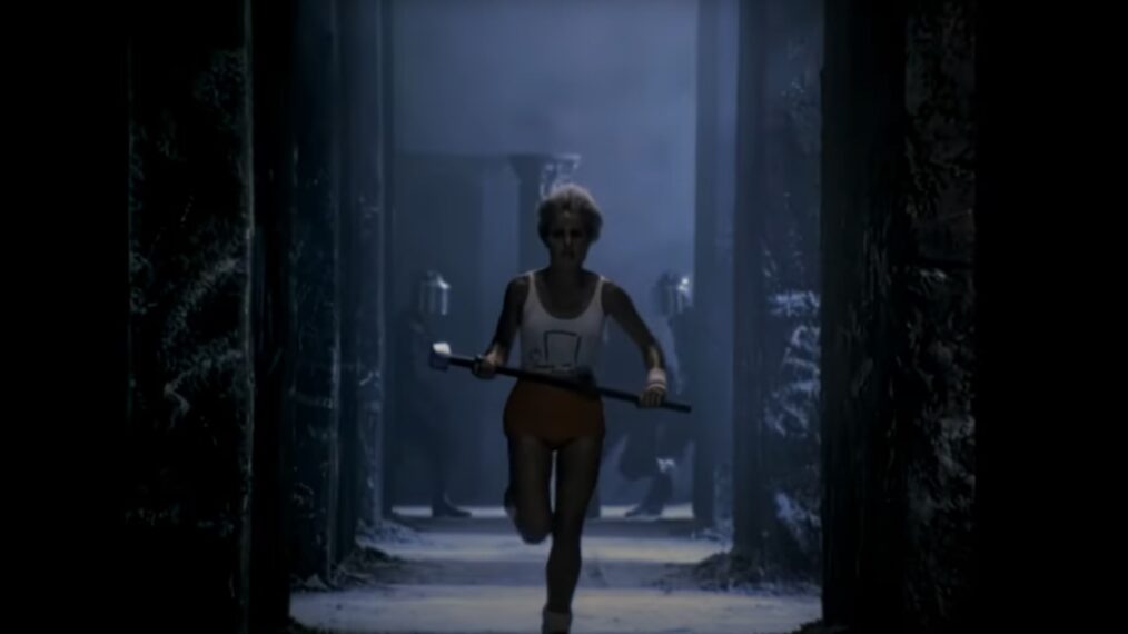image from the commercial for Apple's new Macintosh commercial that aired during the Super Bowl on Jan. 22, 1984. In the center of the image is a blond-haired woman wearing red shorts and a white tank top that has an artistic illustration of the Macintosh computer on it. She has a determined look on her face as she runs down an aisle in a dark, dystopian, '1984'-like world, with helmeted stormtroopers behind her in the distance in pursuit. The woman is clutching a hammer in both hands as she runs, preparing to throw it at a giant screen (not seen here) on which Big Brother is giving a propaganda speech.
