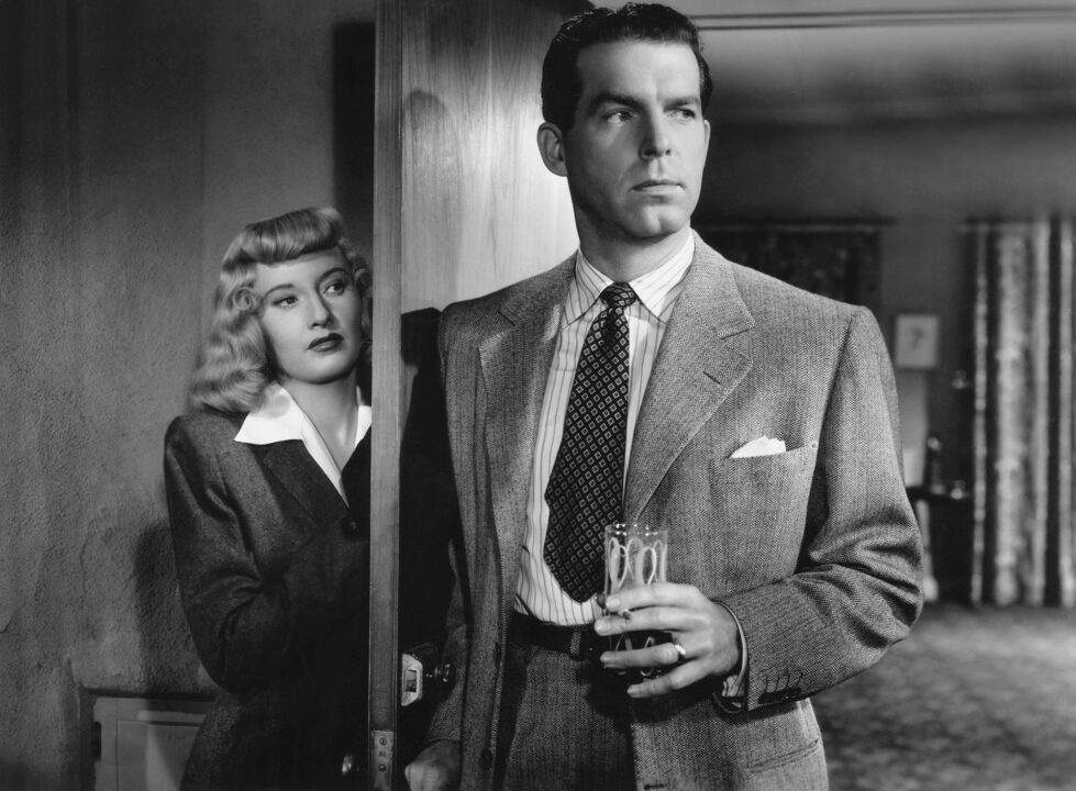 black and white image from the 1944 film "Double Indemnity." On the right of the photo is Fred MacMurray, holding a drink in his right hand and with his left hand in the pocket of his dress pants as he is looking anxiously toward his left after having just entered a room. To the left, hiding behind the room's open door, is Barbara Stanwyck looking at him.