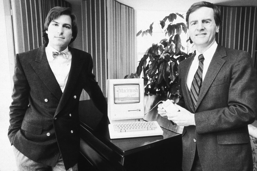black and white image from Jan. 16, 1984, of Steve Jobs (on left of horizontal photo, wearing a white dress shirt, dark dress jacket and striped bow tie, and standing with his right hand in his pants pocket), then chairman of Apple Computers, along with Apple's president, John Sculley (on right of photo wearing a white dress shirt, somewhat lighter dark jacket and longer, standard tie). The men are posing on either side of Apple's new Macintosh computer, which is on a table, its screen reading: "Macintosh Insanely Great." Sculley is holding the computer's mouse in both hands.