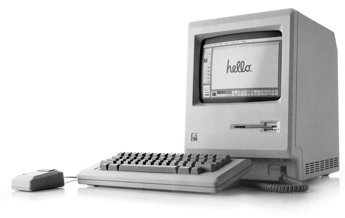 black-and-white image of the first Macintosh computer from Apple in 1984. The monitor of the computer stands on the far right of the horizontal photo; on the screen, written in cursive, is the word "hello." In front of the monitor is the keyboard, and slightly to the left of that, the mouse.