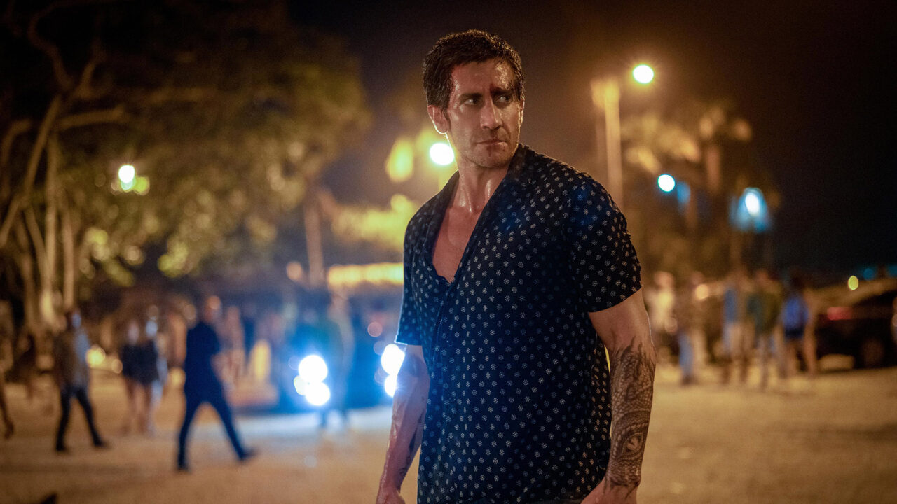 image from the 2024 movie "Road House." It depicts Jake Gyllenhaal as Dalton. He is wearing a casaul shirt that is unbuttoned enough to show some of his chest. He is crossing a street at night and look back a bit behind him with a sense of anger and concern.