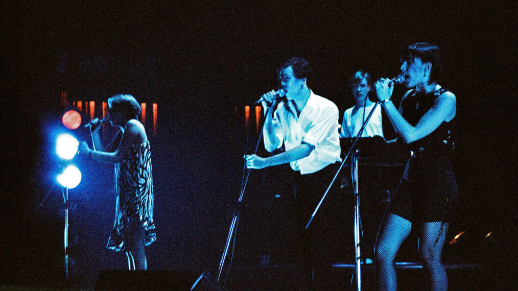 image from a 1981 live performance by The Human League at London's Rainbow Theatre. Most of the image is dark, but the performers onstage are illuminated with blue lighting as they sing. From left are Susan Ann Suylley, wearing a black-and-white striped dress; Philip Oakey, wearing a white dress shirt with sleeves rolled up; and Joanne Catherall, wearing a short black dress. They are all clutching their microphones and microphone stands as they give a passionate performance.