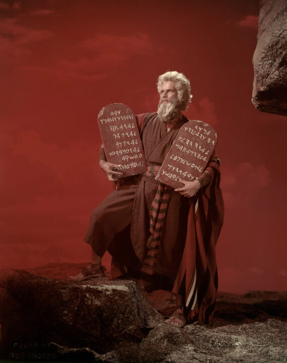 vertical image of Charlton Heston in character as Moses in the 1956 movie "The Ten Commandments." Moses, with greyish-white hair and beard and wearing a red garment and robe, is standing on a cliffside against a red sky background, his right leg up on one higher portion of the cliff and with his left leg lower and supporting him. He is looking up in awe as he holds, one in each arm, two stone tablets contained etched inscriptions of the Ten Commandments, written in ancient Hebrew.