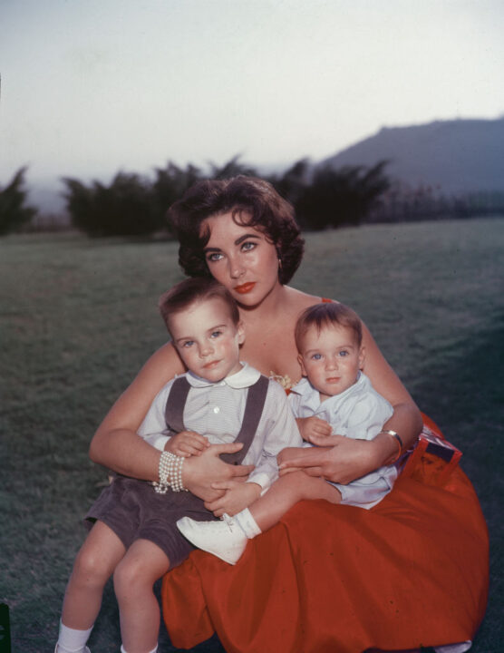British-born actor Elizabeth Taylor sits in a red evening dress with her sons Michael (L) and Christopher Wilding on her lap, mid 1950's