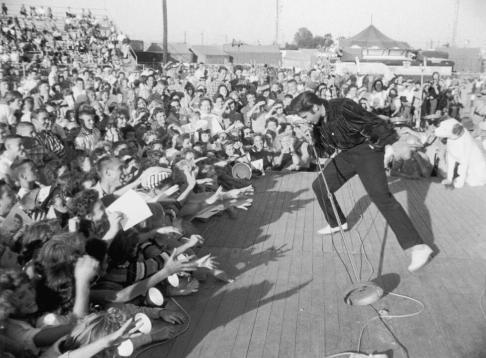 circa 1957: American singer and actor Elvis Presley (1935-1977) performing outdoors on a small stage to the adulation of a young crowd