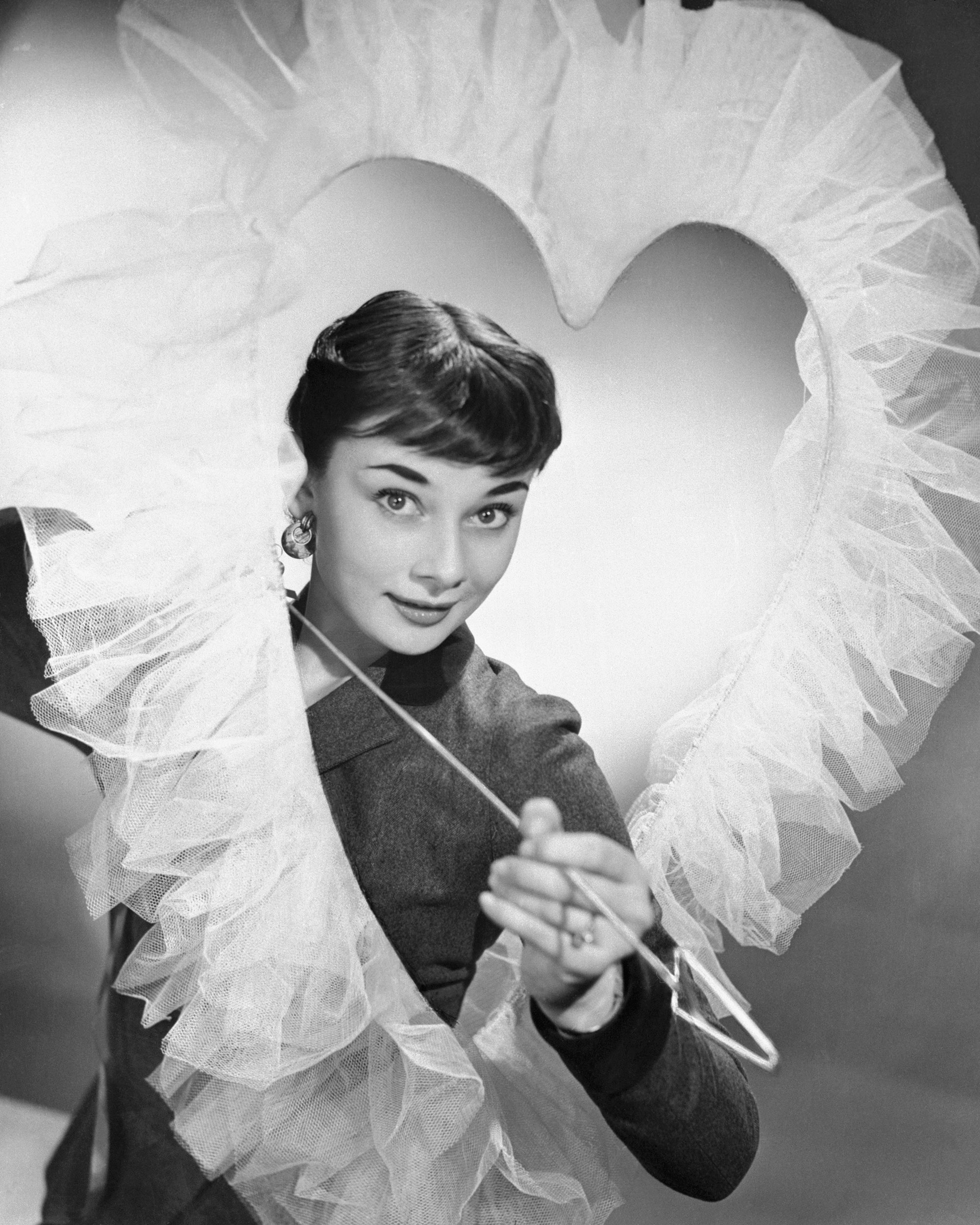 Audrey Hepburn makes a lovely Valentine decoration, complete with Cupid-like smile and arrow as she looks through a large heat-shaped opening with frills around it.