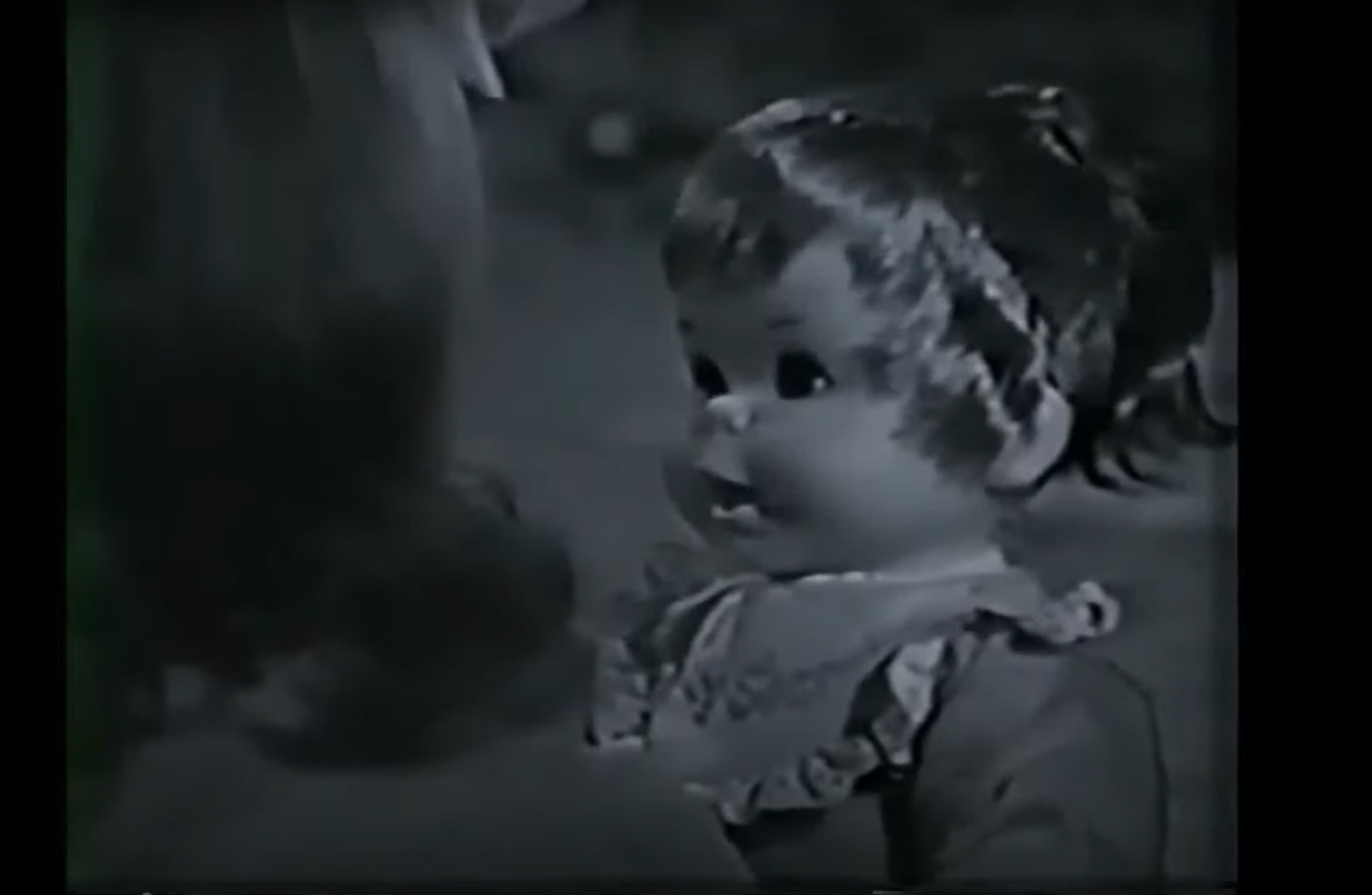 black and white image from a 1966 commercial for Mattel's Baby Secret doll. The doll is on the right and looking at the little girl on the left (seen from the back) who is holding her.