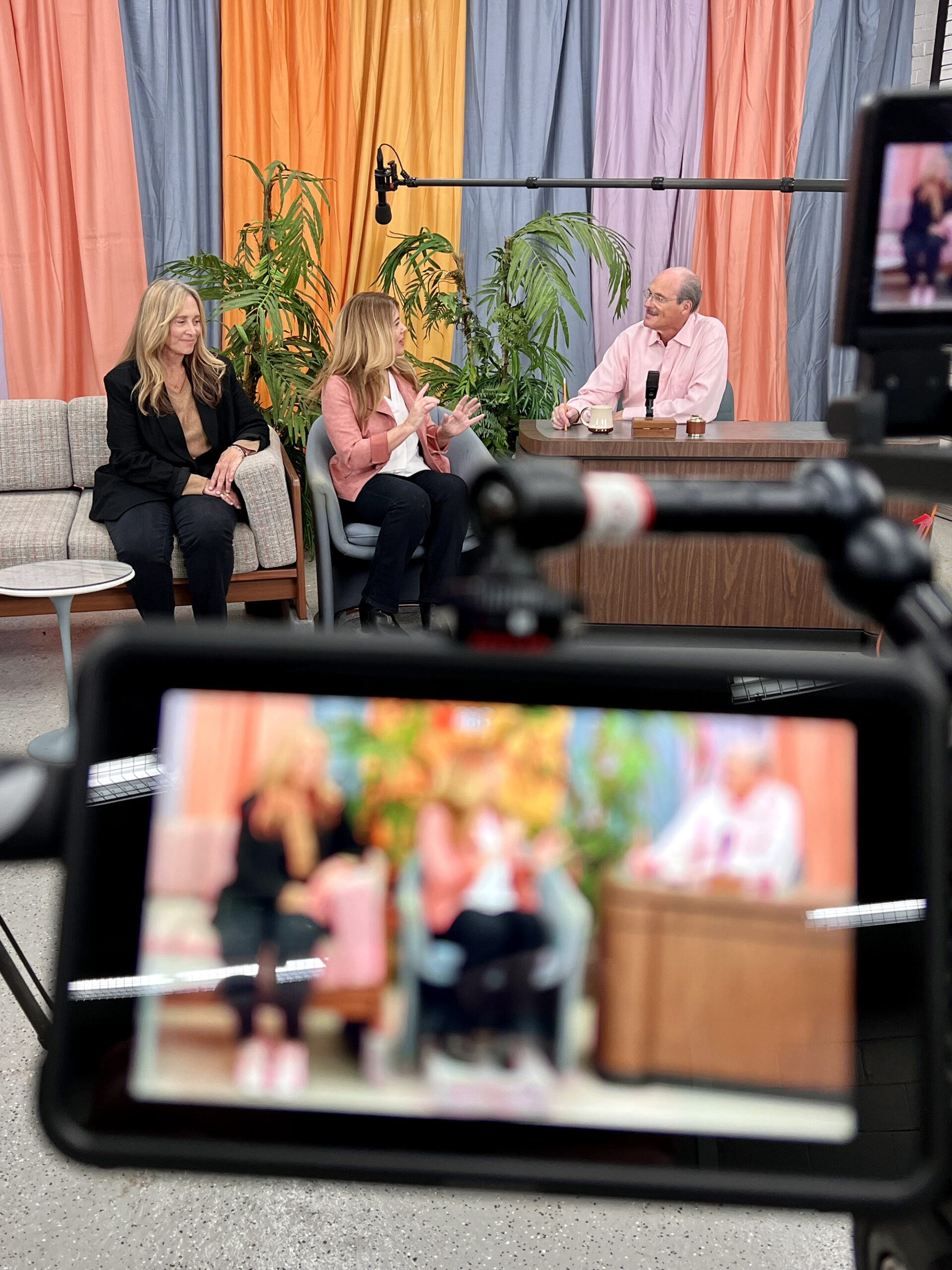 image from the Season 5 premiere of MeTV's "Collector's Call." In the background, seated left-to-right, are expert Marcia Tysseling and host Lisa Whelchel in chairs, and collector Stewart Berkowitz seated behind a desk on the original set of "The Tonight Show Starring Johnny Carson." In the foreground of the image, we are looking at them through the viewfinder of a video camera.