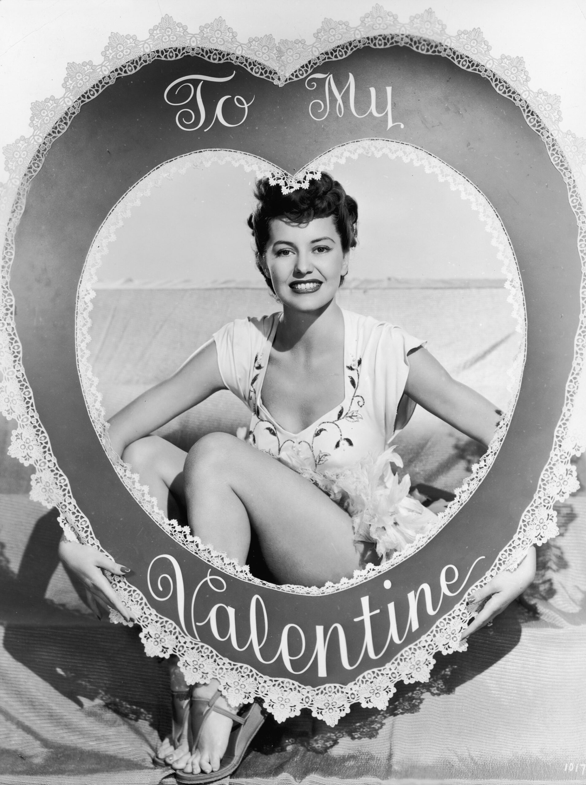 1945 studio portrait of American actor and dancer Cyd Charisse holding and smiling through a large cutout heart valentine that reads, 'To My Valentine.'