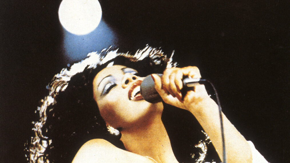 image of Donna Summer performing, circa 1975. The image is a tight shot from her chest up to her head as she holds a microphone in her left hand up close to her mouth. Her head is back and her eyes are closed, revealing light blue eye shadow, as she passionately sings into the mic. Above and behind her, a circular spotlight shines down.