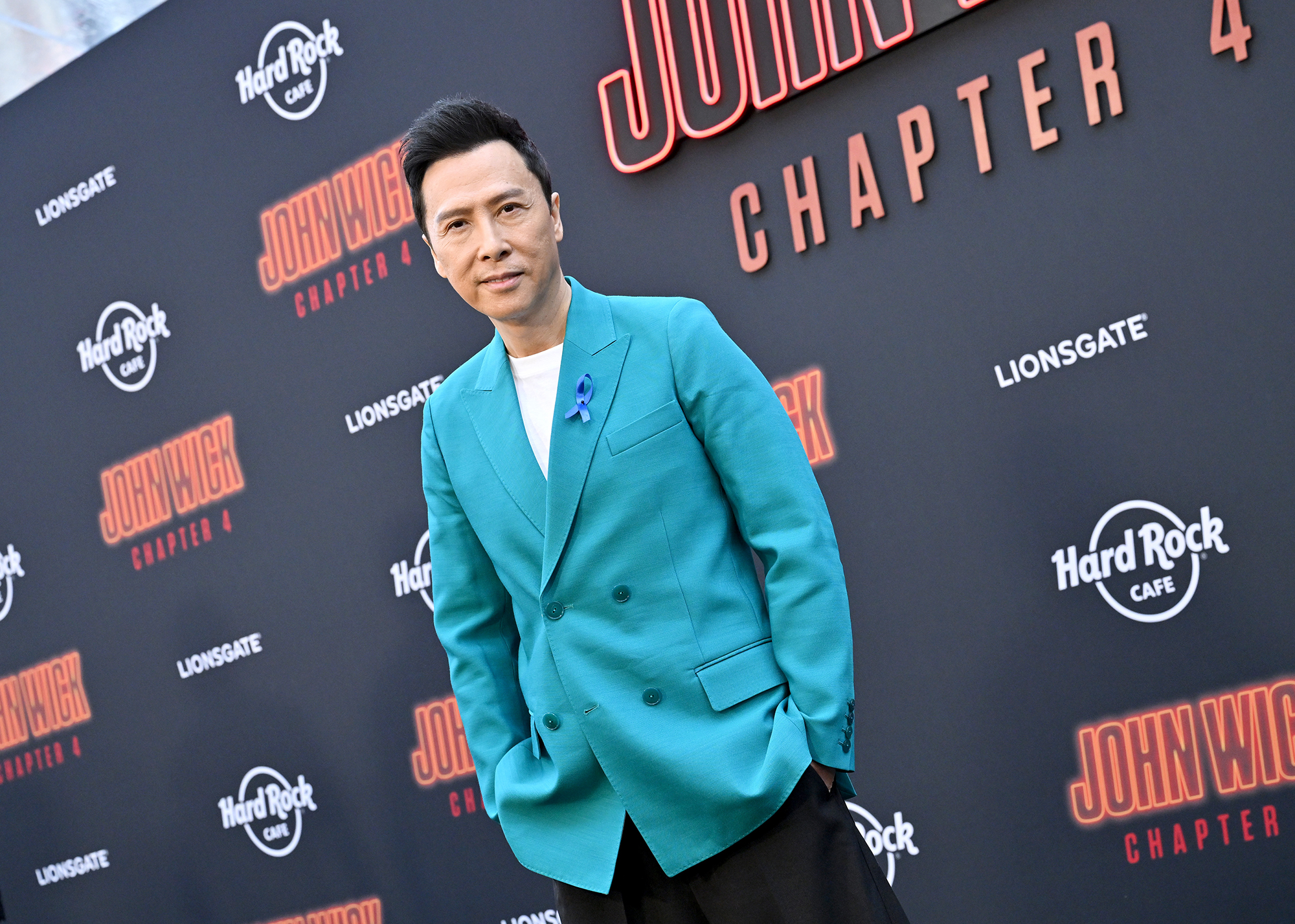 celebrity image of martial arts star/actor Donnie Yen outside of the March 2023 premiere for "John Wick: Chapter 4." Yen is wearing a white shirt underneath an aqua-colored jacket, with his hands in the pockets of his dark-colored pants as he poses for a shot on the red carpet.