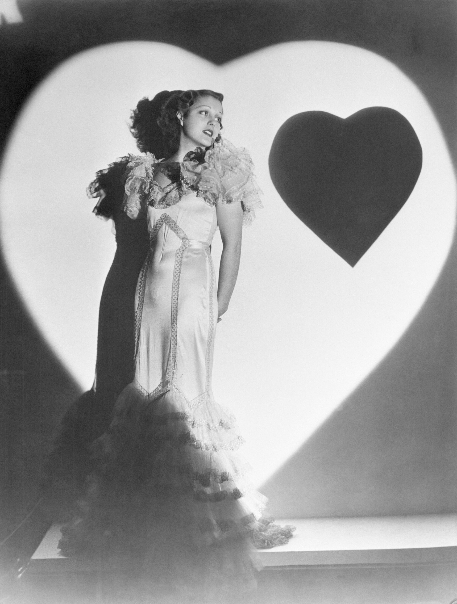 Actress Frances Drake, wearing a fancy gown with frills at the bottom and at the shoulders, leans against a wall that is illuminated by a heart-shaped spotlight, and with a paper heart affixed to the wall next to her, in this black-and-white Valentine's Day studio portrait from the early 1930s.