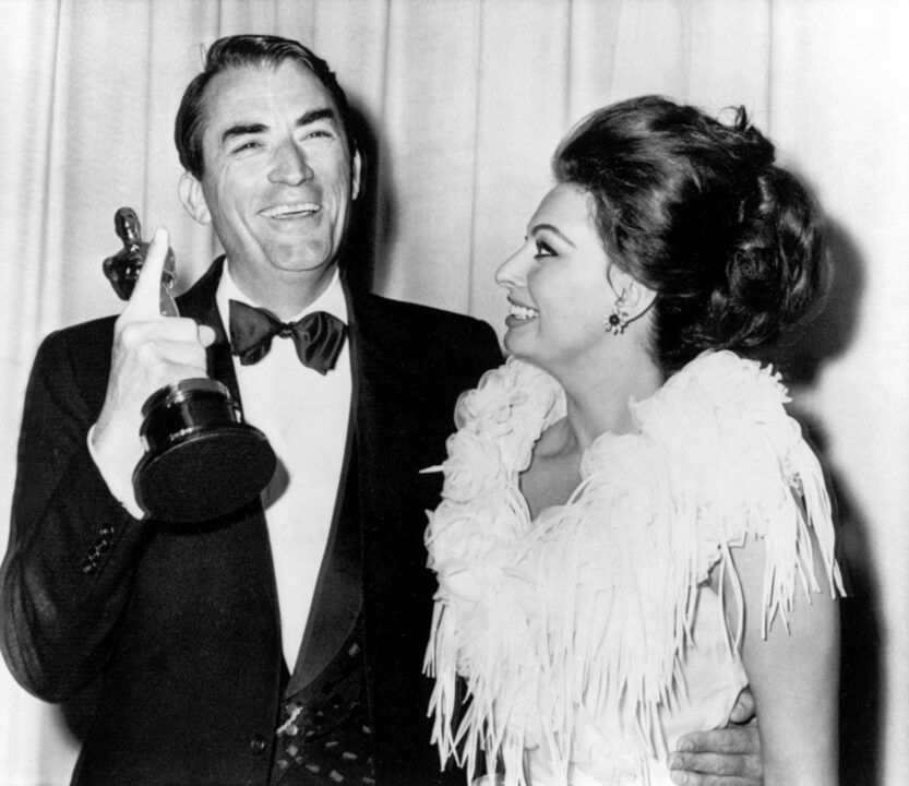 black-and-white image from the 35th Academy Awards. Best Actor winner Gregory Peck (To Kill a Mockingbird), wearing a tuxedo and smiling, is on the left of the photo, holding the Oscar he has just won in his right hand. His left arm is around Sophia Loren, also smiling and wearing a white formal dress, who had won Best Actress the previous year and presented Peck with his award.