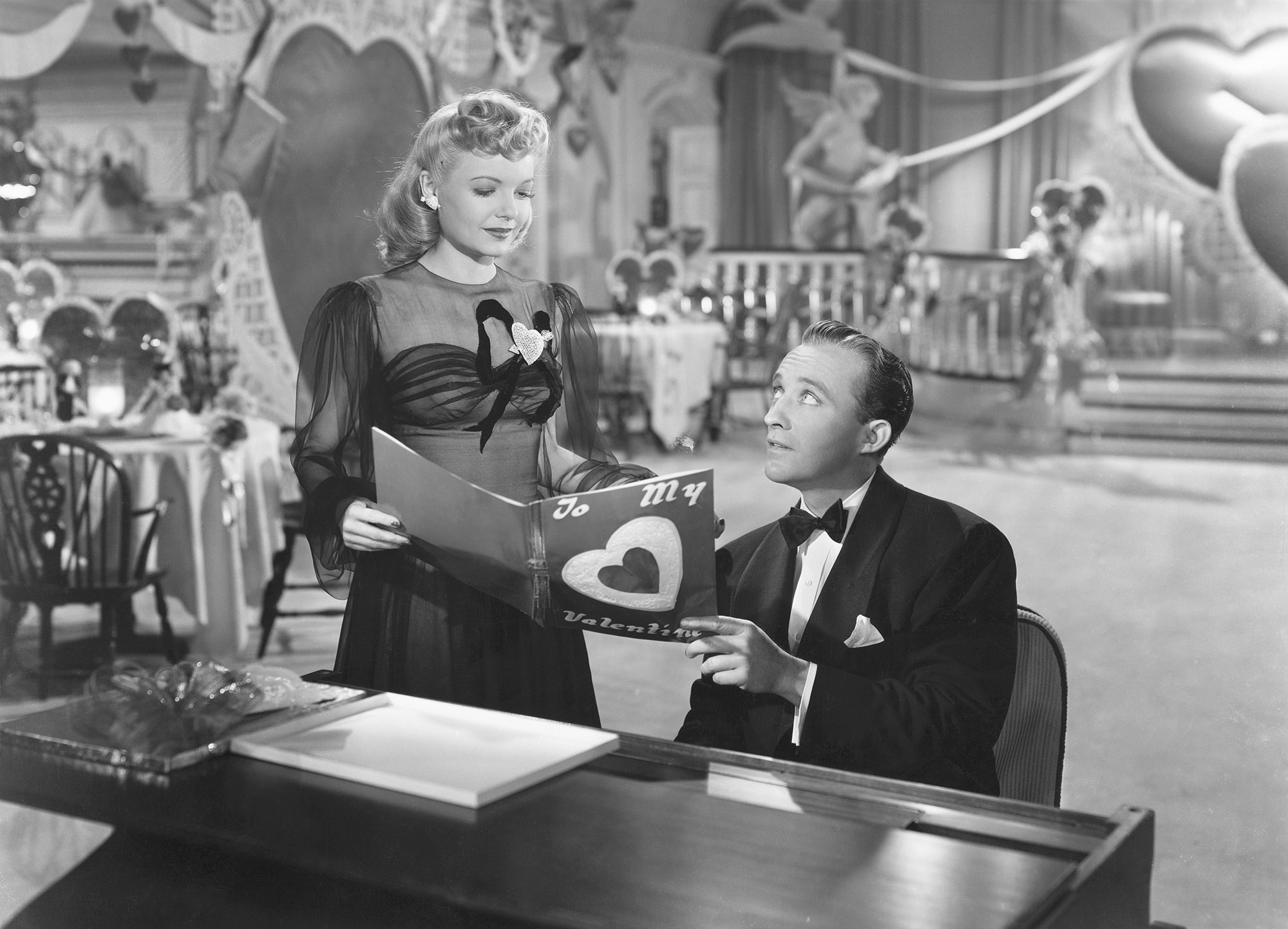 black and white image from the 1942 movie "Holiday Inn." Bing Crosby is seated at a piano, wearing a tuxedo and looking up at Marjorie Reynolds, standing next to him and wearing a fancy dress. They are both holding a Valentine's Day card, and the set behind them is decorated with hearts for the holiday.