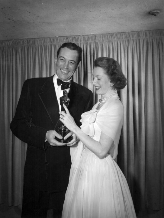 black and white photo from the 1949 Oscars ceremony. Deborah Kerr, on the right wearing a white formal dress, hands the Best Original Screenplay Oscar statuette to John Huston, smiling and wearing a tux, for "The Treasure of the Sierra Madre."