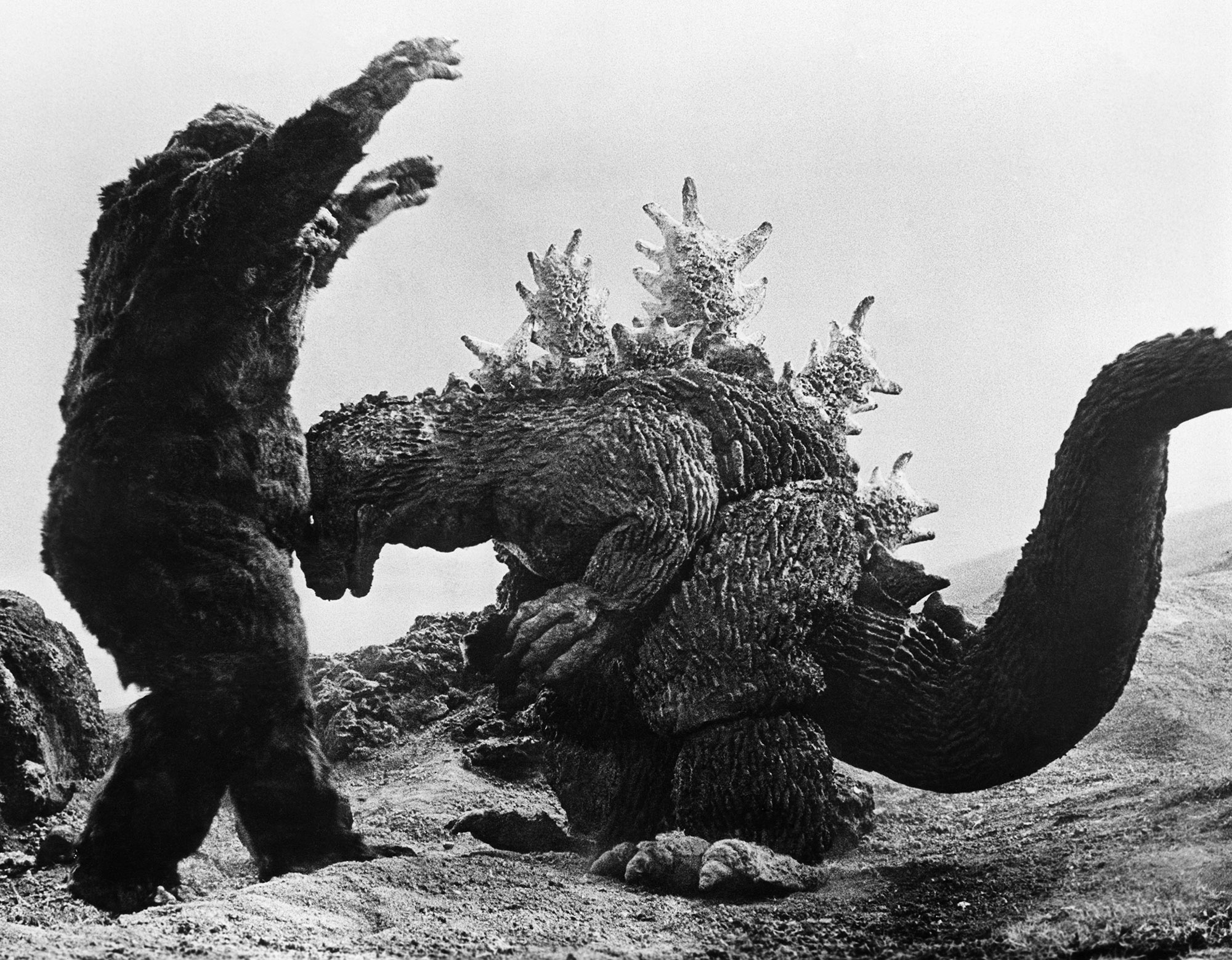 black and white still from the 1962 movie "King Kong vs. Godzilla." The title monsters are fighting, with Kong on the left, standing with his arms in the air as he is being knocked back by Godzilla, who is bent forward and head-butting Kong in the chest.
