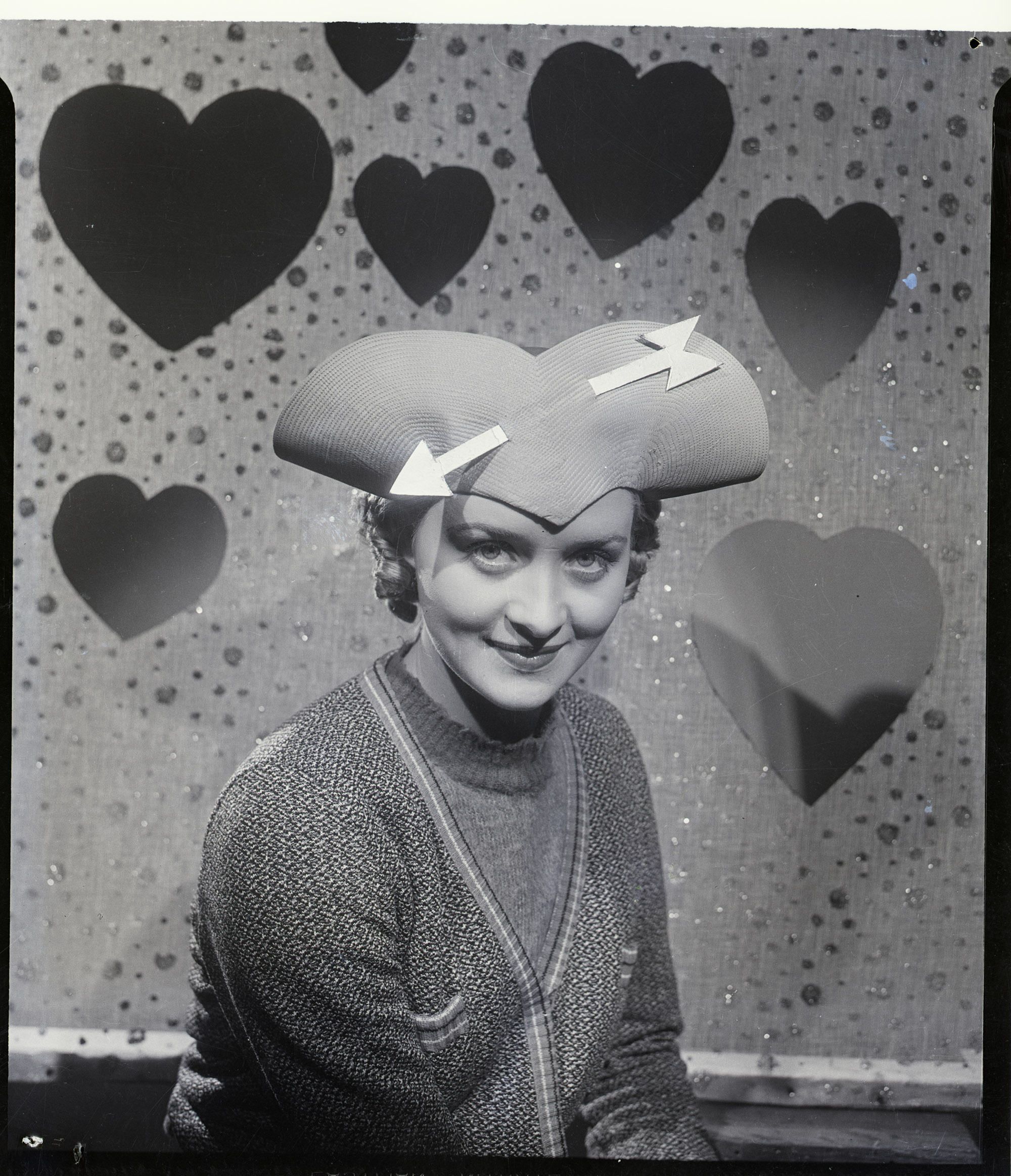 a "Be my valentine" hat is worn by actress Marian Marsh. The hat has curves in the shape of a heart to fit on the head with a gilded arrow, piercing the head gear to form a decoration. Marsh is seated wearing the hat against a backround of hearts for Valentine's Day.