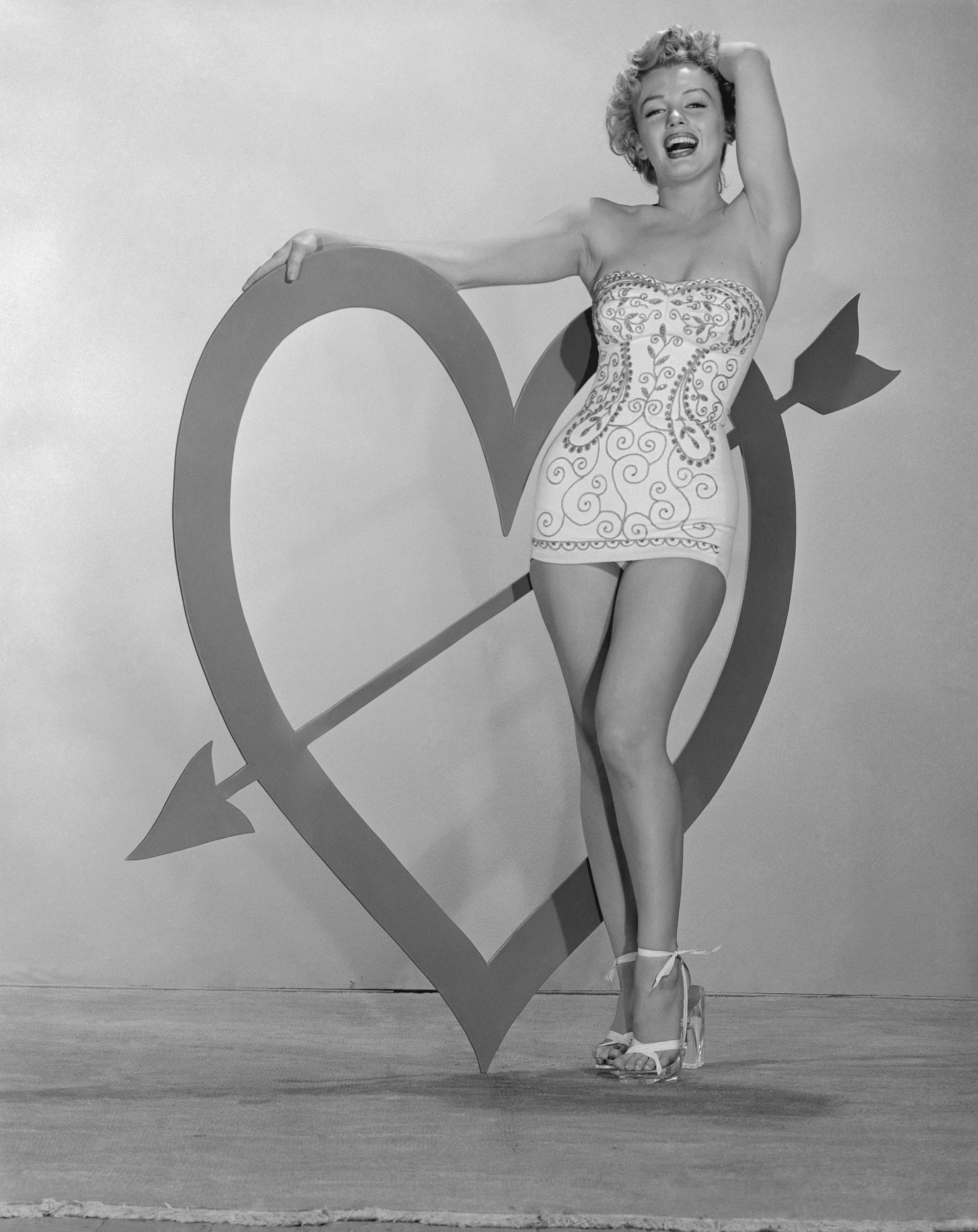 Marilyn Monroe stands beside a large heart shape with an arrow through it for Valentine's Day in a 1955 studio portrait