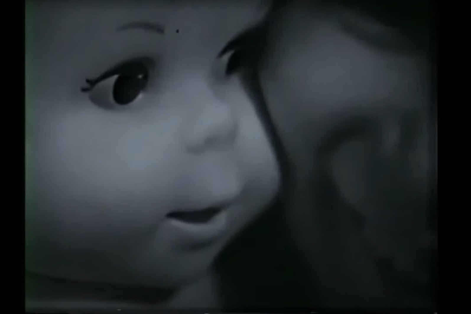 black and white screengrab from a 1966 commercial for Mattel's "Baby Secret" doll. It is an extreme closeup with the doll's face on the left, turned toward the voice of its human owner (played by young Eve Plumb). The baby doll appears to be whispering into the girl's ear, and the girl is smiling.