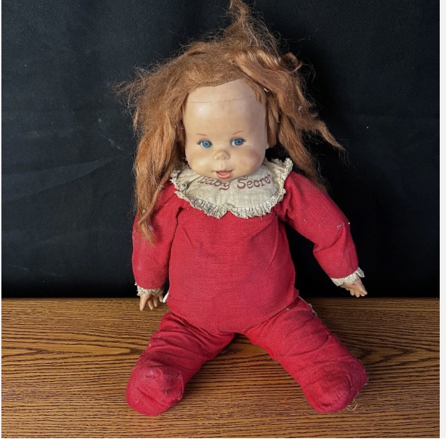 color image of a used and very distressed/dirty 1960s Baby Secret doll from Mattel, for sale on eBay. The doll is seated on a table, wearing a red onesie with a white collar on which reads "Baby Secret" in red letters. The doll has red scraggly and thinned-with-age hair.