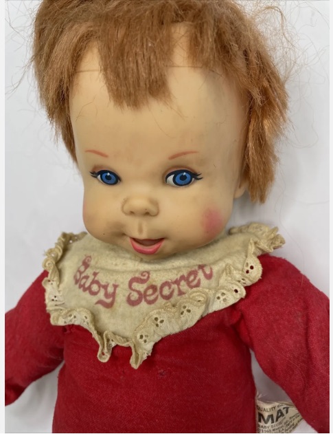 color image of a 1960s used and very distressed Mattel "Baby Secret" doll for sale on eBay. The doll is dirty and worn, and is wearing a red onesie with a white collar on which reads: "Baby Secret" in red letters. The doll has red hair which has thinned with age.