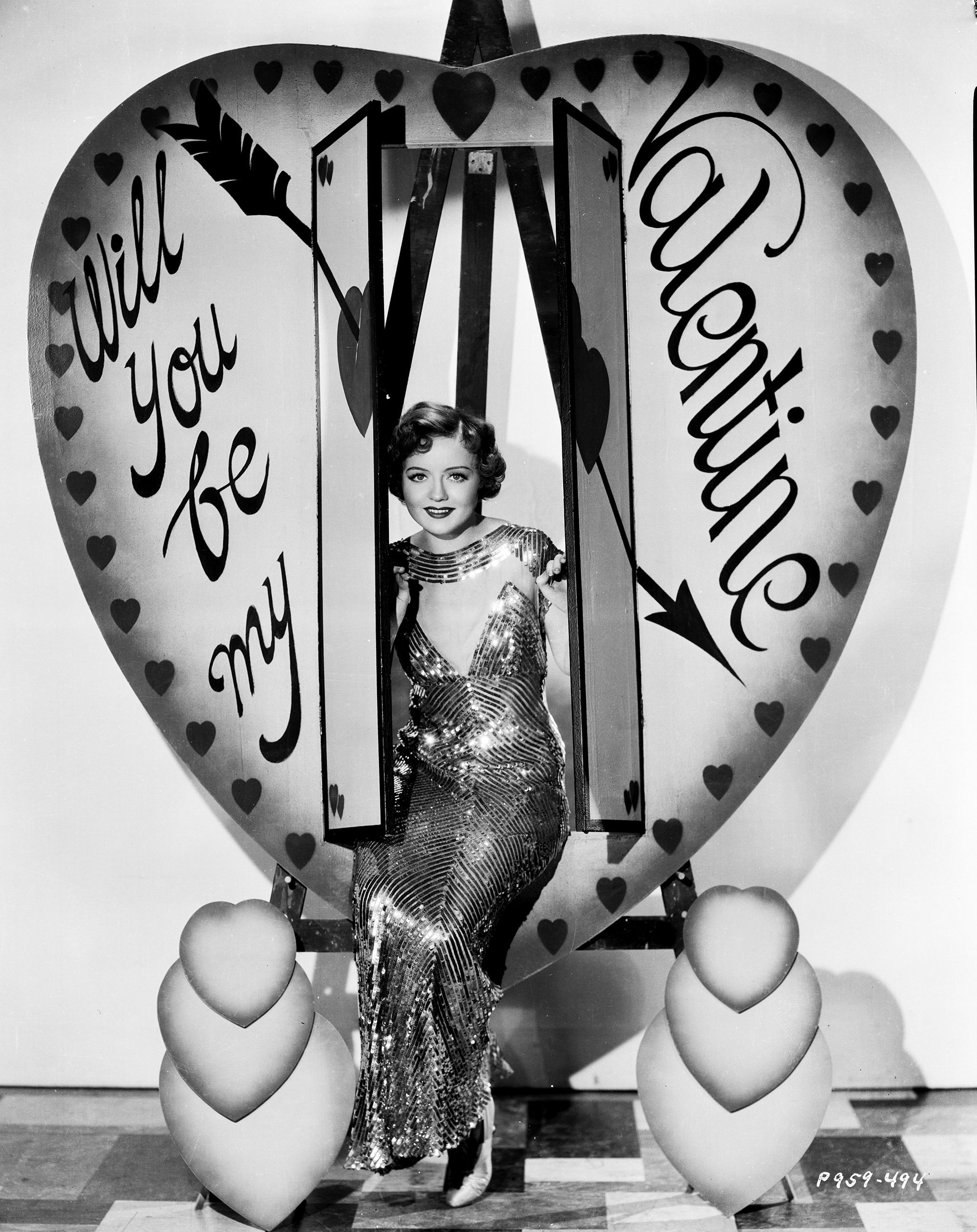 Portrait of actress Nancy Carroll sitting inside a prop heart labeled 'Will You Be My Valentine', for Paramount Pictures, 1929.