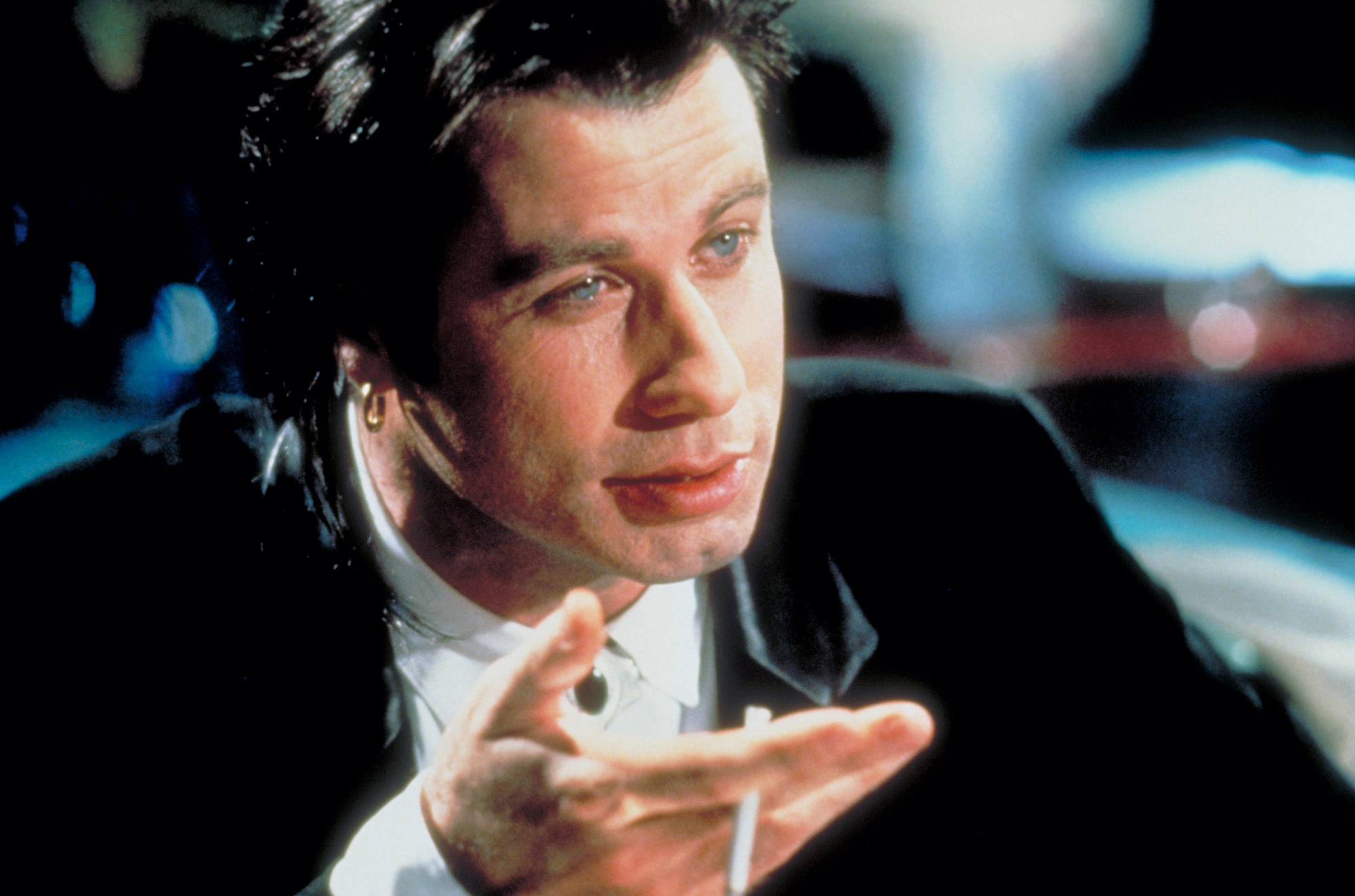 image from the 1994 movie "Pulp Fiction." it is an extreme closeup of John Travolta as Vincent Vega, wearing a dark suit and holding a cigarette in his right hand as he talks to someone across from him.