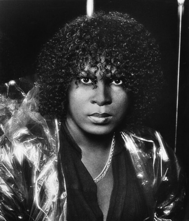 black and white portrait of disco singer Sylvester (Sylvester James Jr.) circa 1978. He is a Black man with dark curly hair, wearing a black shirt that is opened to reveal his smooth bare chest and a chain he's wearing around his neck. He is a somewhat intent expression on his face as he looks ahead at the camera.