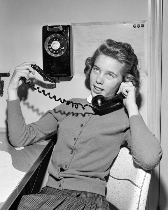 black and white image of a teenage girl in the 1950s talking on a rotary dial phone. She is sitting in front of the phone, which is mounted on a wall, and has part of the coiled cord connecting the phone's handset to the base in her right hand while holding the phone to her ear with her left.She is smiling as she listens to the person on the other end of the line.