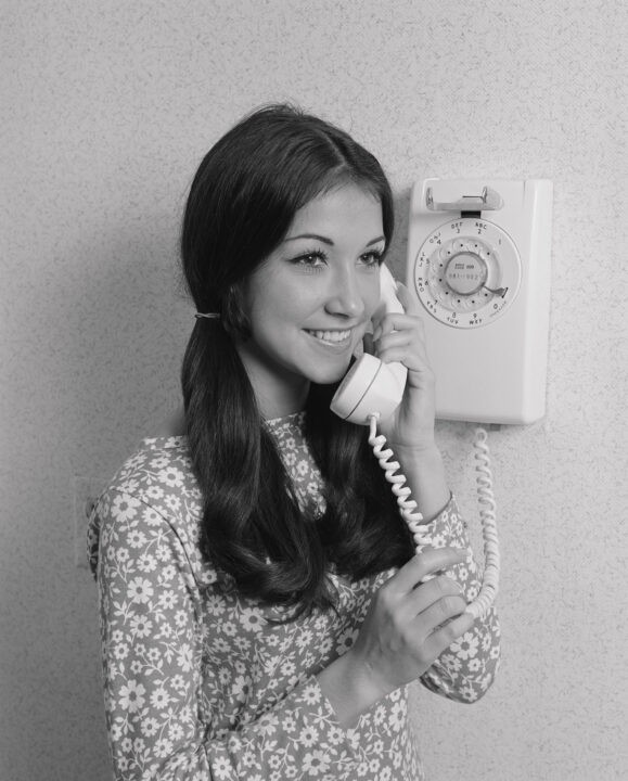 black and white stock image of a teenage girl talking on a rotary dial phone circa late 1960s/early 70s. She has dark hair tied in ponytails on each side and is wearing a lighter-colored dressed with a dotted pattern. She is smiling as she listens while holding the phone up to her ear with her right hand, while standing next to the base of the phone, which is mounted on the wall next to her.