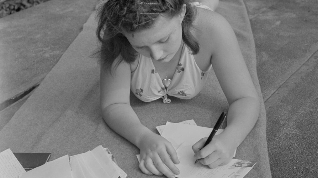 black and white image of a teenage girl writing a letter to her boyfriend in 1942. She is lying on her stomach on a blanket outside on some grass as she write this, wearing a 1940s bathing suit, with some open books in front of her.