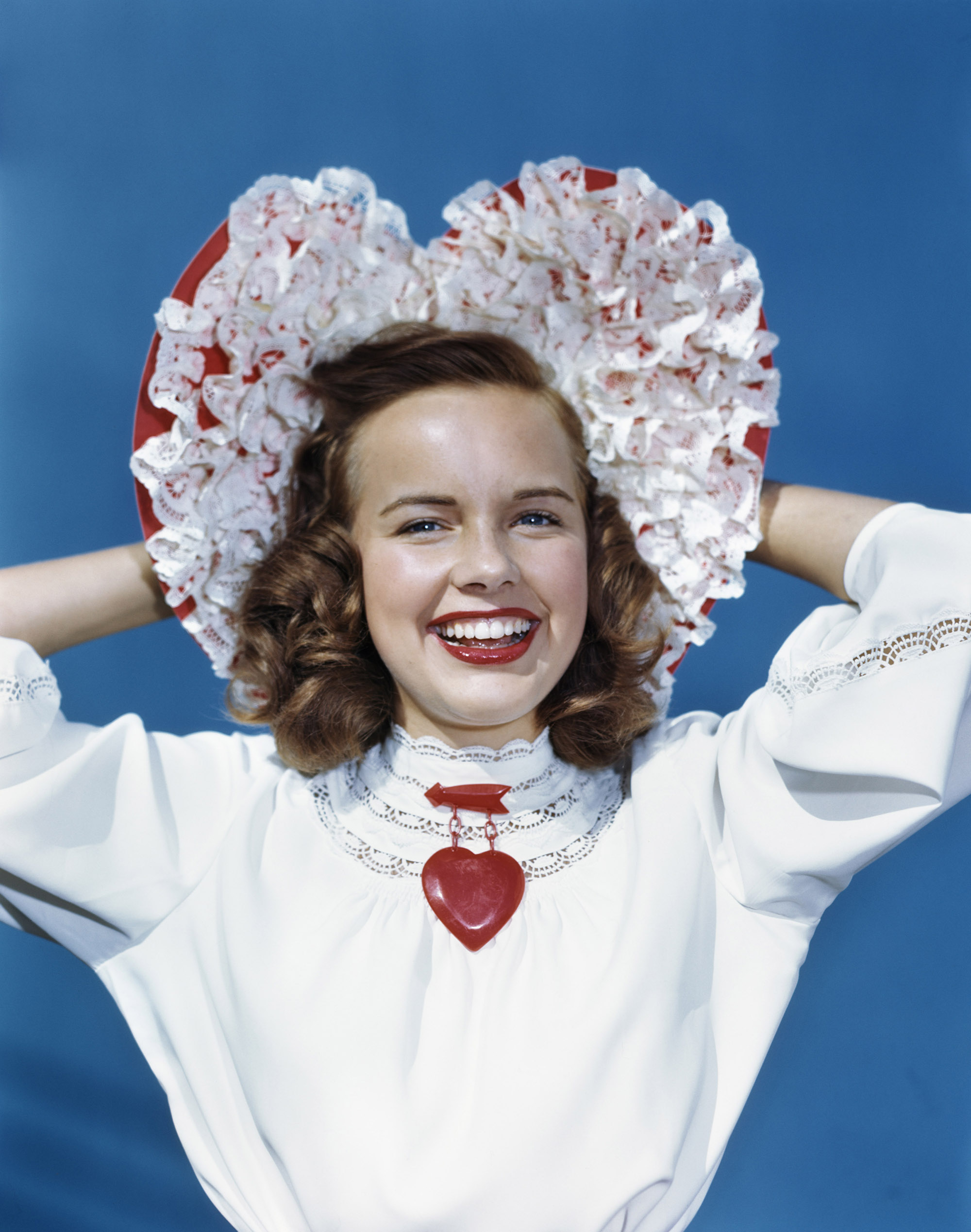 Actress Terry Moore, standing against a light blue background, wears a white blouse, red heart brooch and a white-and-red heart-shaped bonnet as she smiles in a 1949 Valentine's Day studio portrait.
