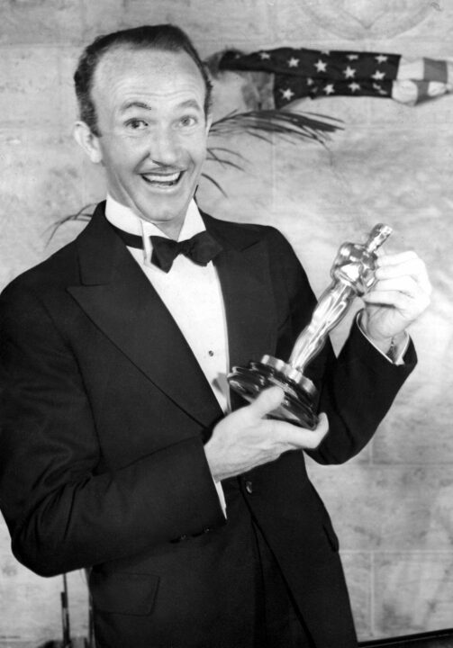 black and white image of the 9th Academy Awards on March 4, 1937. Walter Brennan, wearing a tuxedo, smiles proudly as he poses with the Best Supporting Actor Oscar he won for 1936's "Come and Get It." This was the first year that category was awarded.