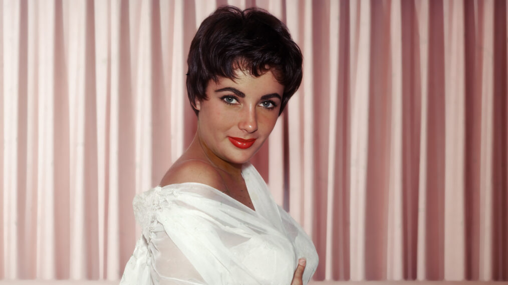 British-born actor Elizabeth Taylor kneels on a bed and wears a sheer white nightgown and robe, circa 1950s.