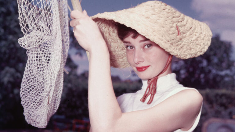 Portrait of Belgian-born American actress Audrey Hepburn (1929 - 1993) as she wears a peculiar hat and sleeveless blouse and holds a pool cleaning net beside a dry swimming pool, early 1950s