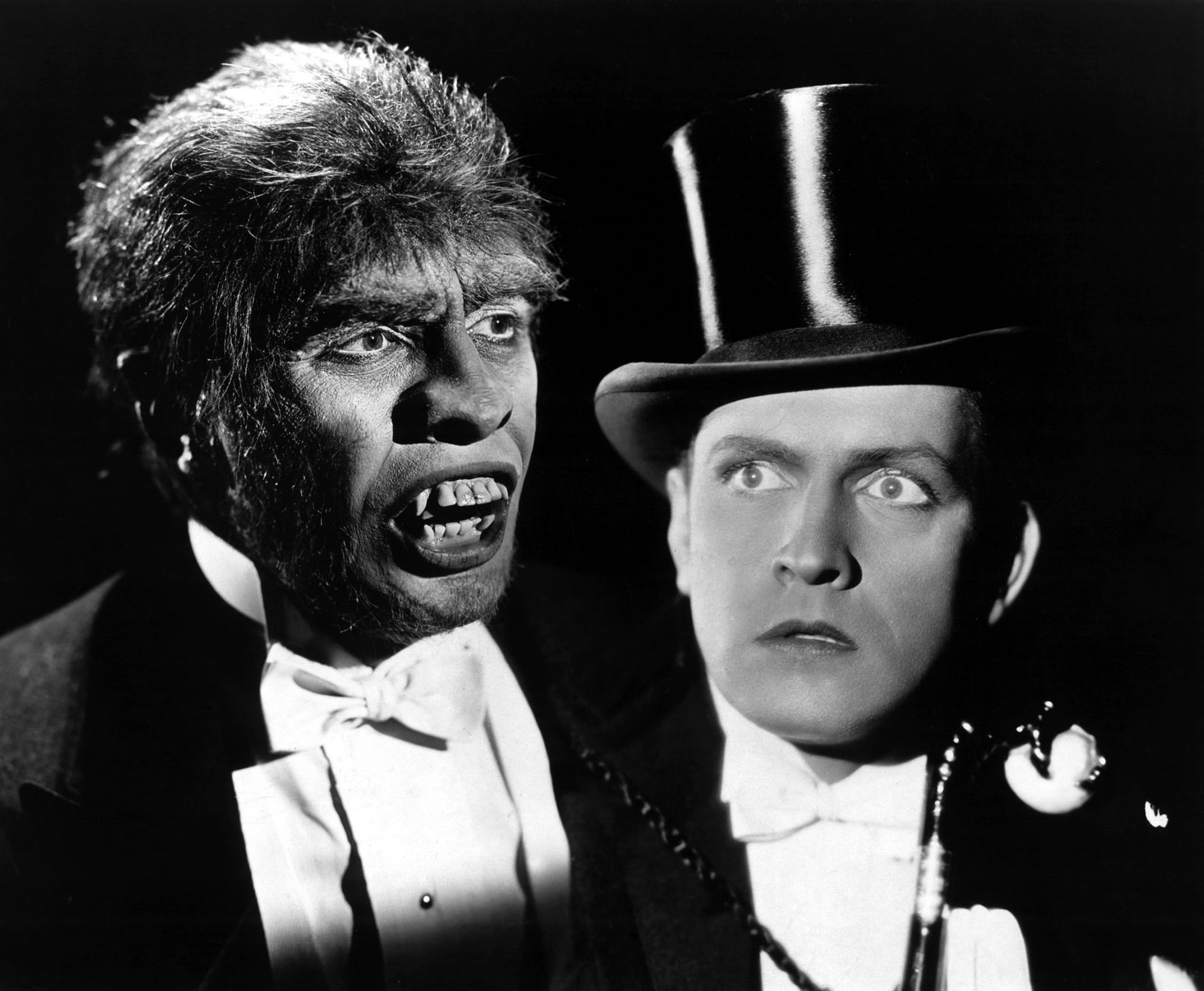 black and white promotional image for the 1931 movie "Dr. Jekyll and Mr. Hyde," showing star in dual role as Mr. Hyde (on the left, a deformed monstrous looking creature wearing a dress shirt and tux) and Dr. Jekyll, on the right, looking in shock at Mr. Hyde while also wearing a tux and shirt, with a top hat.
