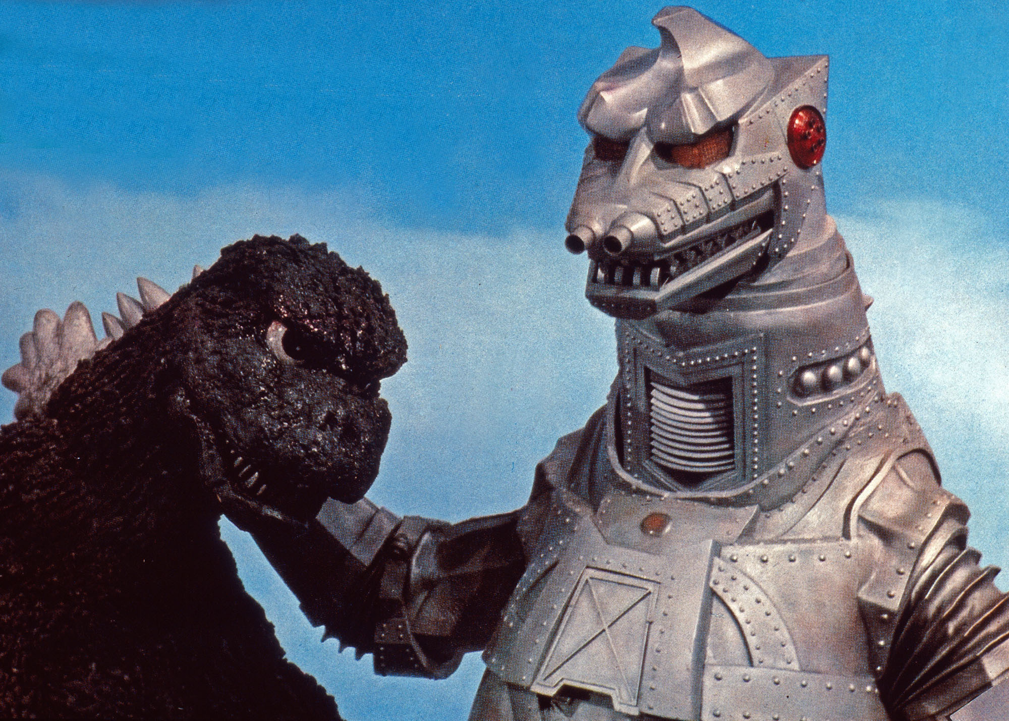image from the 1974 movie "Godzilla vs. Mechagodzilla." It features closeup headshots of Godzilla, on the left, and Mechagodzilla, on the right, grappling with each other against a blue sky background.