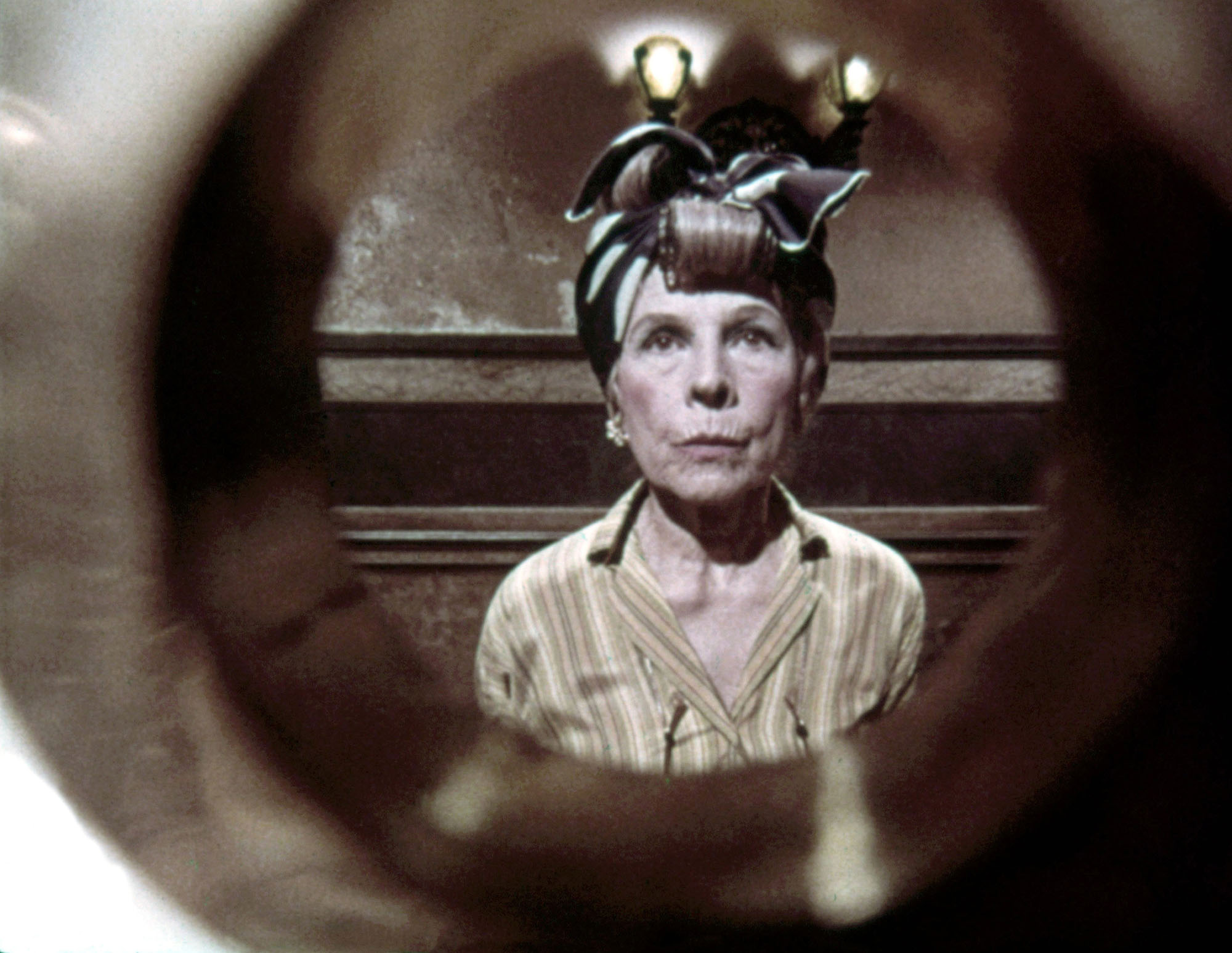 image from the 1968 movie "Rosemary's Baby." We are looking through the peephole of an apartment door at Ruth Gordon's character, Minnie Castevet, who is standing outside the door waiting for it to be opened. She is frowning and looks disturbed by something as she waits, and has her hair up in a wrap.