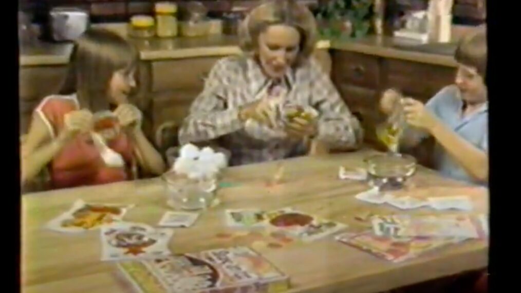 image from a 1981 commercial for the Shake-an-Egg Easter egg dye coloring kit. A mother is seated at a table in between her daughter, on the viewer's left, and son, on the right. They have the Shake-an-Egg kit open and spread out on the table, and they are placing eggs in bags filled with coloring crystals to shake them.