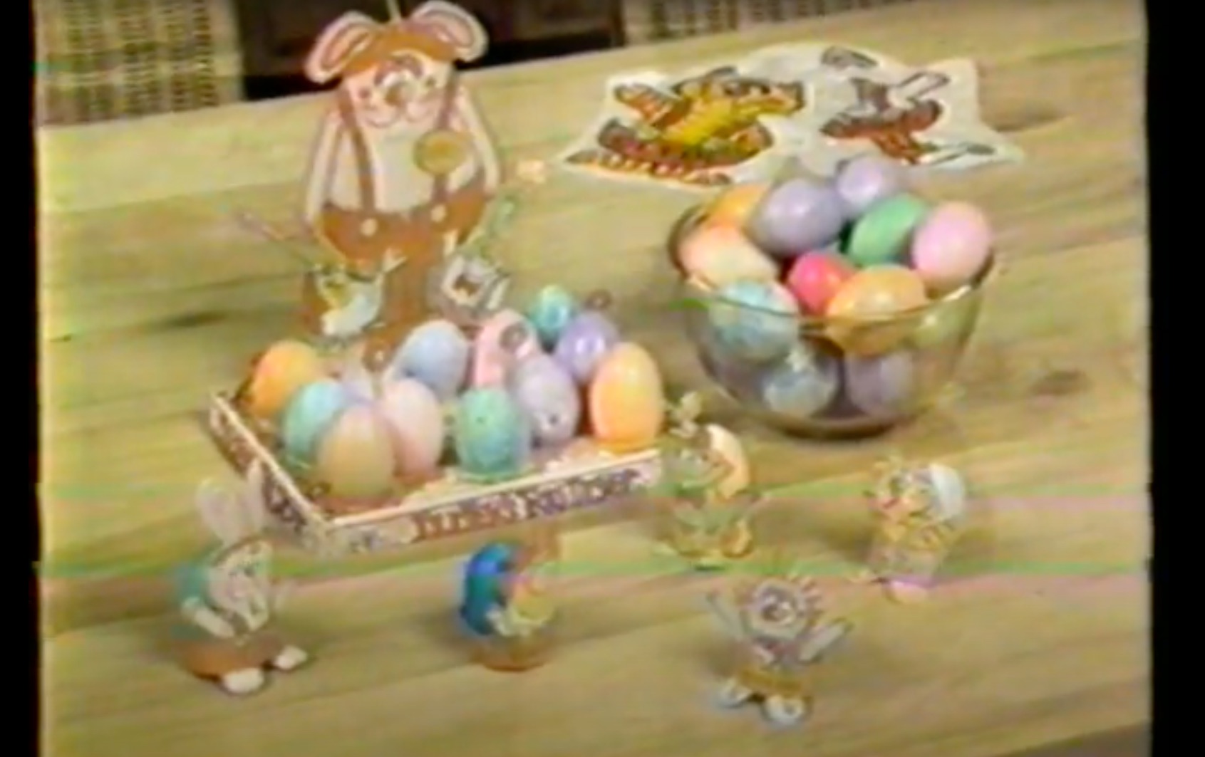 image from a 1981 commercial for Shake-an-Egg Easter egg coloring kit. On a table is a bowl of colored eggs. Next to it is a paper stand with Dudley Rabbit, the Shake-an-Egg mascot, looking over more colored eggs that are placed in the paper stand. In front of that, more eggs are being held by paper cutouts of Dudley and other characters.