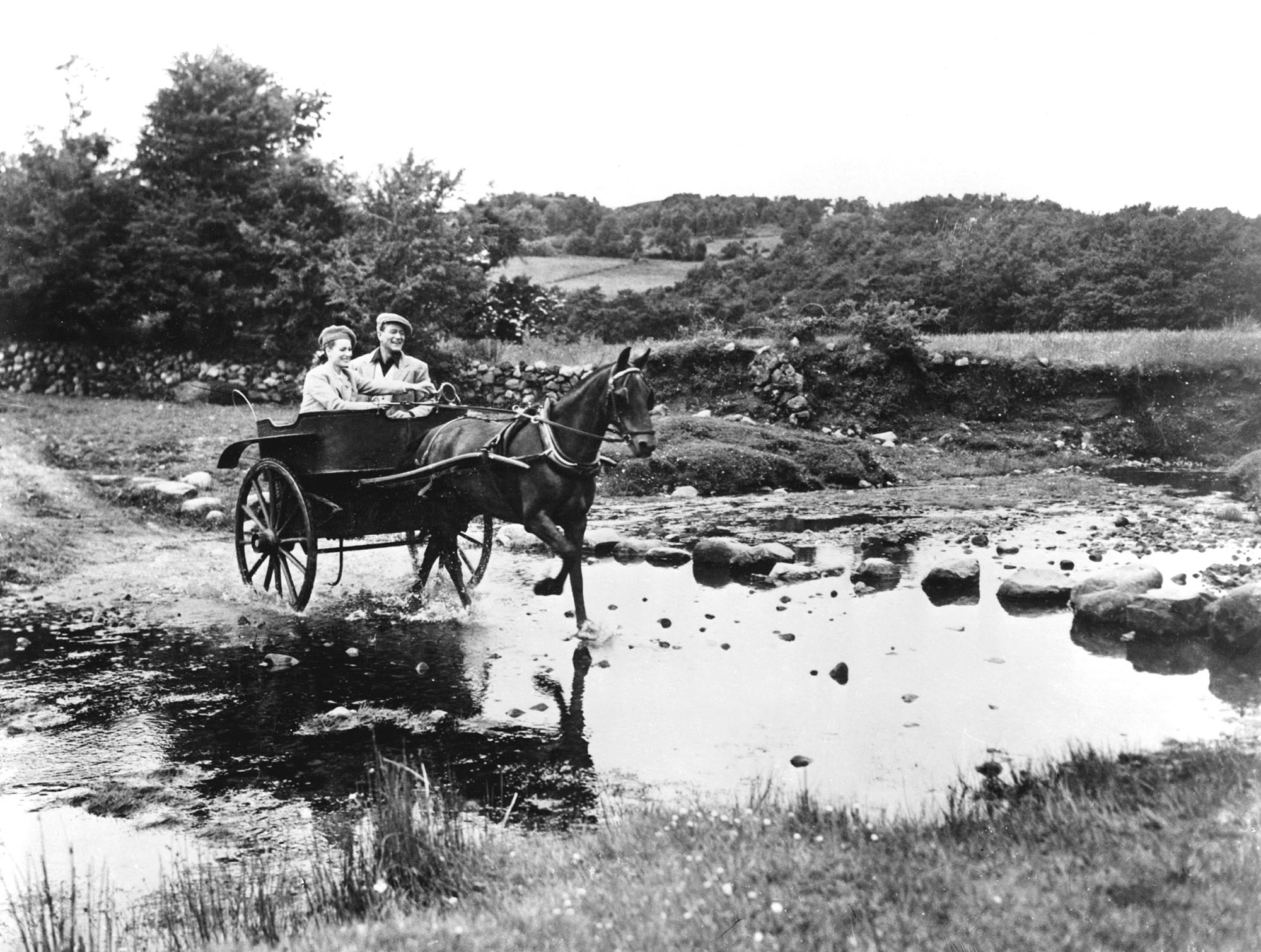 black-and-white image from the 1952 movie "The Quiet Man." It depicts stars Maureen O'Hara and John Wayne riding in a horse-drawn wagon across a pond in the Irish countryside.