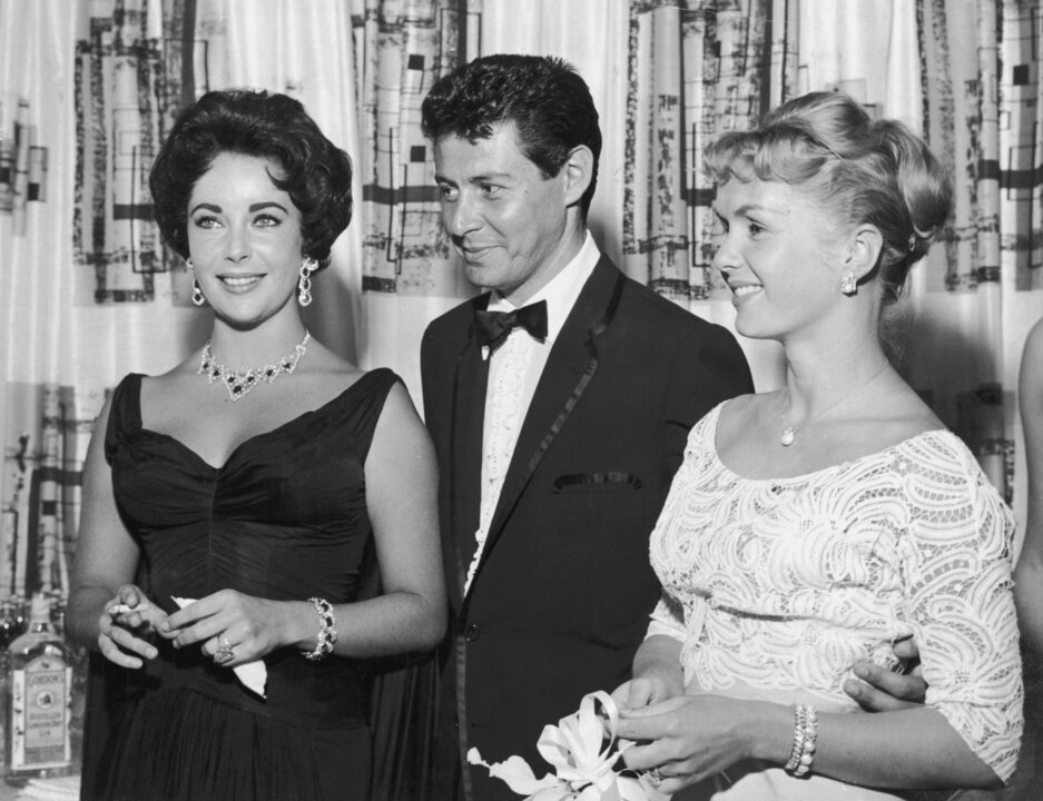 1958: American singer Eddie Fisher, wearing a tuxedo, stands with arm around his wife, American actor Debbie Reynolds (R) and smiles while looking at British-born actor Elizabeth Taylor, smoking a cigarette, Las Vegas, Nevada. The next year Fisher left Reynolds and married Taylor. 