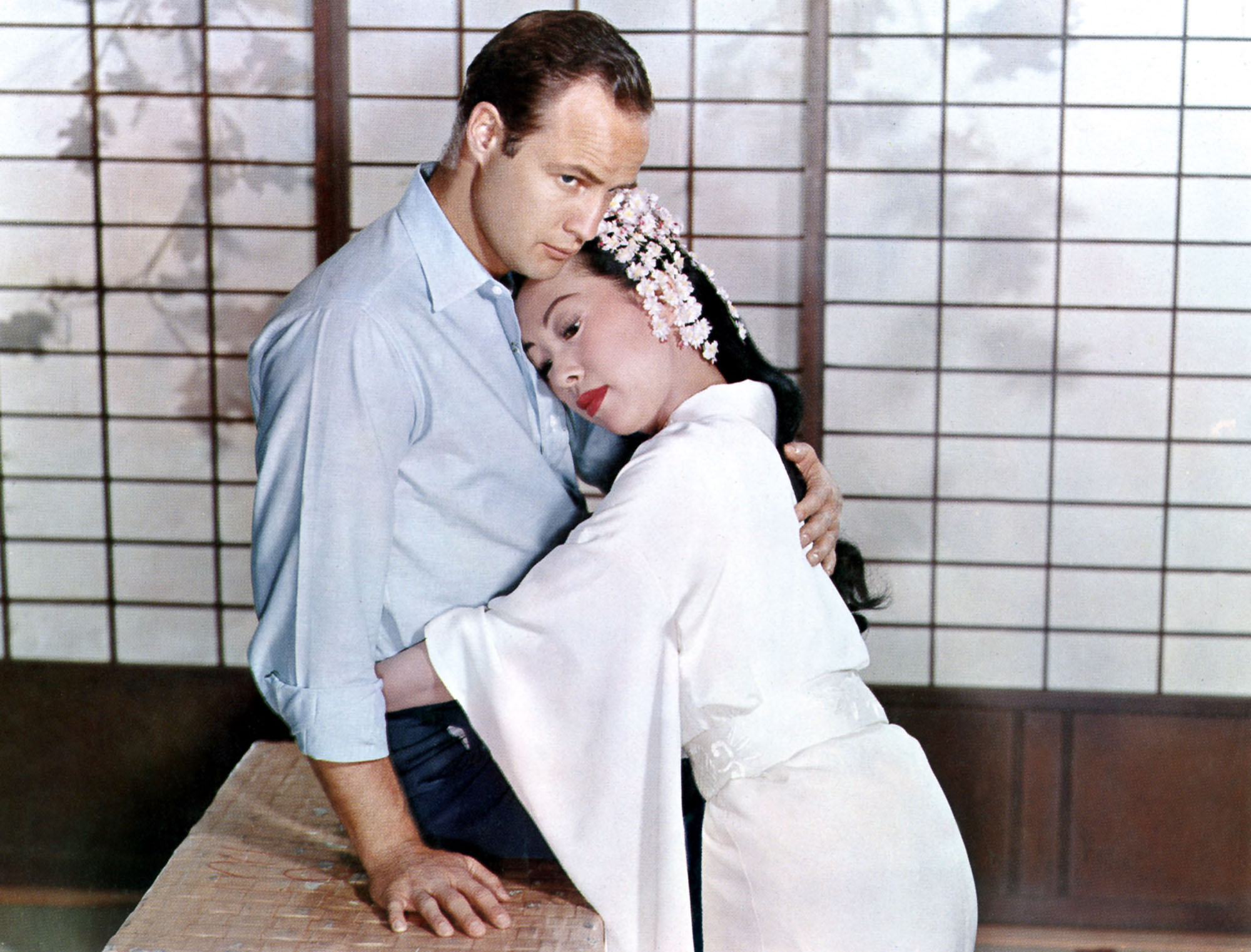 image from the 1957 movie "Sayonara." Marlon Brando's character is on the left, seated in a traditional Japanese room. He is embracing Miiko Taka's character, on the left; she is also seated, wearing a white robe and is returning the embrace. 