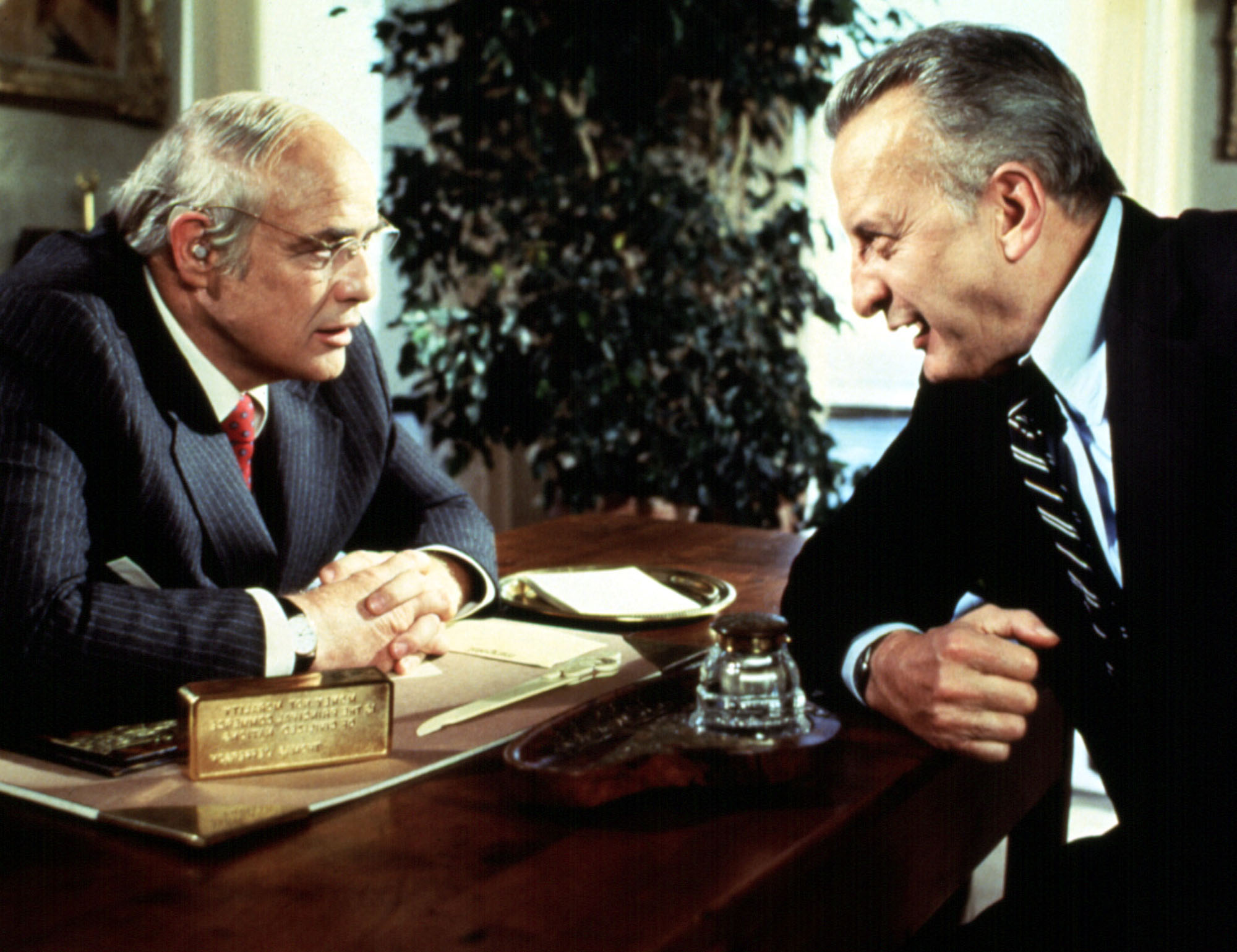 image from the 1980 film "The Formula." On the left, seated behind a desk, is Marlon Brando. Sitting just in front of him on the other side of the desk is George C. Scott. Their characters are leaned on the desk closing in toward each other, as if in argument.