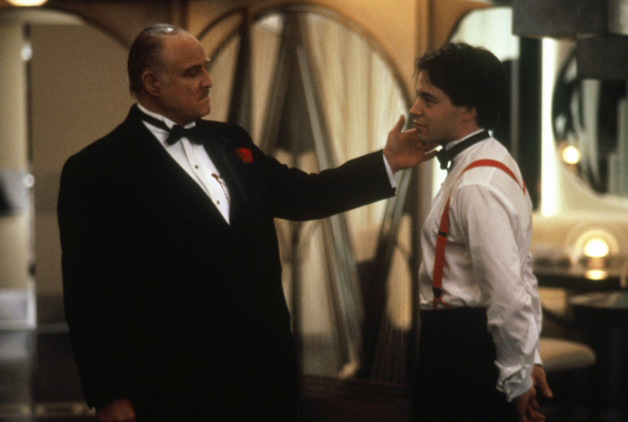 image from the 1990 movie "The Freshman." On the left is Marlon Brando, wearing a tuxedo. He is reachout and touching the cheek of Matthew Broderick's character, on the right and facing Brando and wearing a tuxedo shirt (his jacket is off).